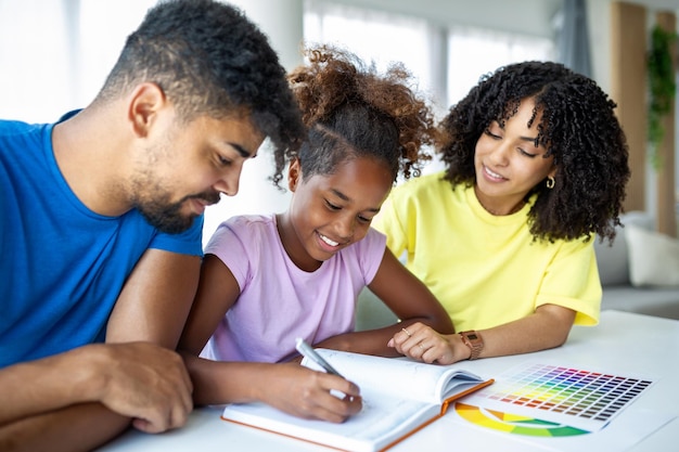 Vorderansicht von afroamerikanischen Eltern, die ihrer Tochter bei den Hausaufgaben am Tisch helfen Foto eines jungen Mädchens, das von ihren Eltern zu Hause unterrichtet wird