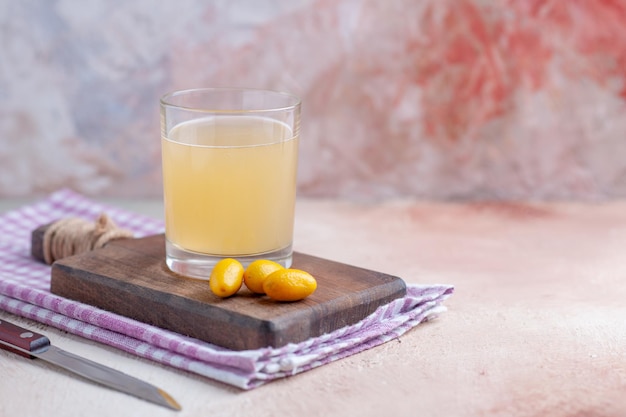 Vorderansicht von Fruchtsaft und Kumquats auf Holzschneidebrett auf lila gestreiftem Handtuch auf der rechten Seite auf pastellfarbenem Hintergrund