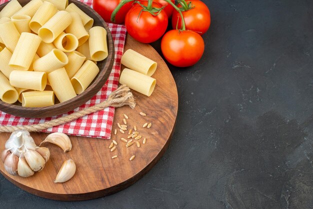 Vorderansicht von rohen Nudeln innerhalb und außerhalb einer braunen Schüssel auf rot gestreiftem Handtuch Knoblauchreis auf rundem Holzbrett Tomatenseil auf der rechten Seite auf schwarzem Hintergrund