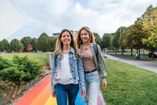 Vorderansicht von zwei fröhlichen Frauen, die auf einem Weg gehen, der mit den Farben des Regenbogens bemalt ist, während sie in die Kamera schauen.