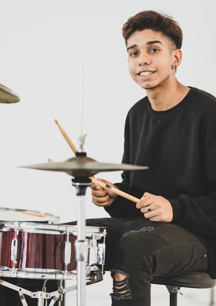 Vorderansichtporträtaufnahme eines jugendlichen Schlagzeugers, der die Musik spielt. Junger Musiker, der die Trommel spielt, während er in die Kamera schaut und lächelt. Juniorstudent, der ein Instrument mit weißem Hintergrund spielt