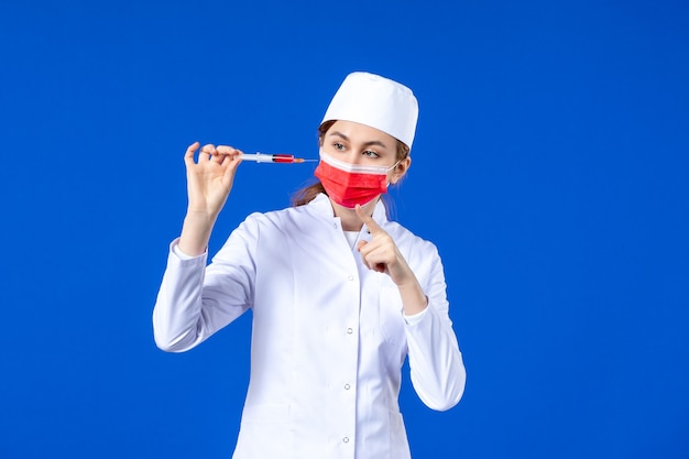 Vordere Ansicht weibliche Krankenschwester im weißen medizinischen Anzug mit roter Maske und Injektion in ihren Händen auf blau