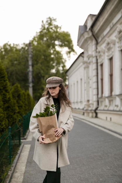 Vorderes Porträt einer stilvollen jungen Frau mit einer Papierpackung der grünen Blattpflanze auf Stadtstadt. Frau, die im kalten Tag geht. Mode, Lifestyle-Konzept.