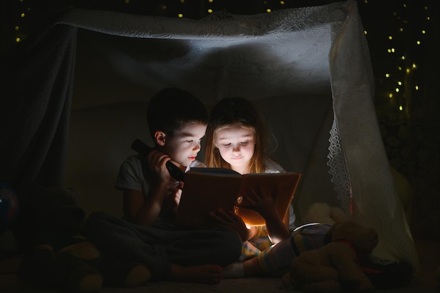 Vorlesen und Familienspiele im Kinderzelt. Junge und Mädchen mit Buch und Taschenlampe vor dem Schlafengehen