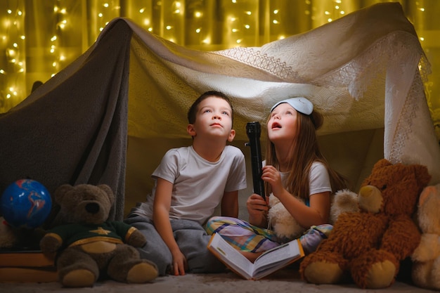 Vorlesen und Familienspiele im Kinderzelt. Junge und Mädchen mit Buch und Taschenlampe vor dem Schlafengehen