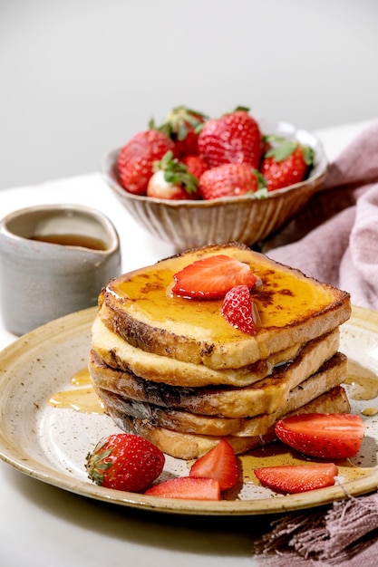 Vorrat an French Toast mit frischen Erdbeeren auf Keramikplatte, Ahornsirup in Keramikkanne und rosa Stoffserviette über weißem Tisch. Frühstück zu Hause