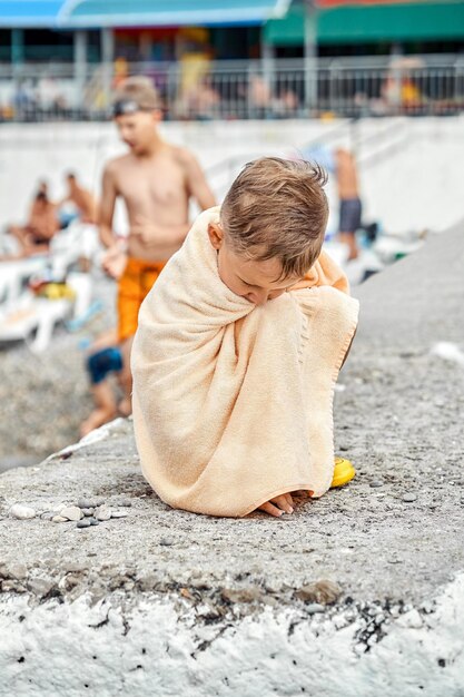 Vorschuljunge, bedeckt mit beigefarbenem Handtuch, sitzt auf dem Pier und wird kalt, nachdem er im Meer geschwommen ist