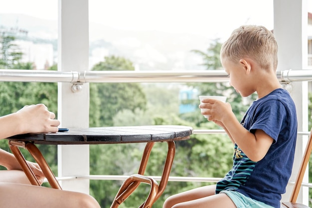 Vorschuljunge trinkt warme Milch aus einem Becher, der am frühen Morgen auf dem Hotelbalkon auf einem Stuhl sitzt