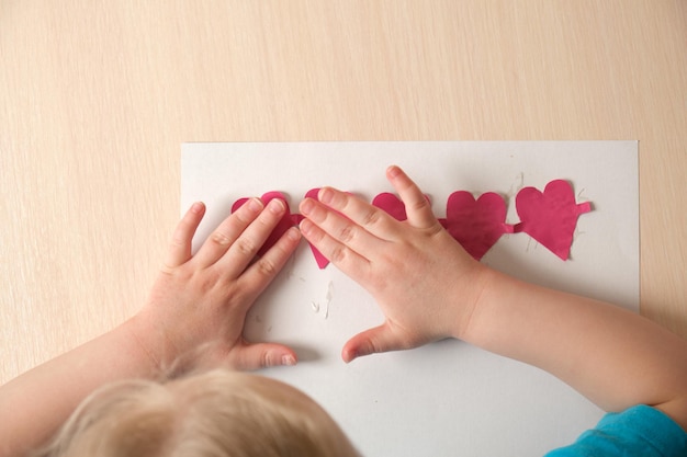 Vorschulmädchen in blauem T-Shirt macht am Tisch sitzende Applikationen, aus farbigem Bougue ausgeschnittene Herzen für die Karte zum Valentinstag, Kreativität der Kinder, atopische Dermatitis an den Händen eines Kindes