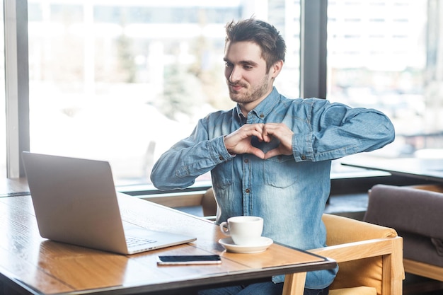 Vos amo! retrato de romântico feliz jovem namorado na camisa azul jeans estão sentados no café e mostrando a sua namorada uma forma de coração com os dedos através de uma webcam online. interior, dentro