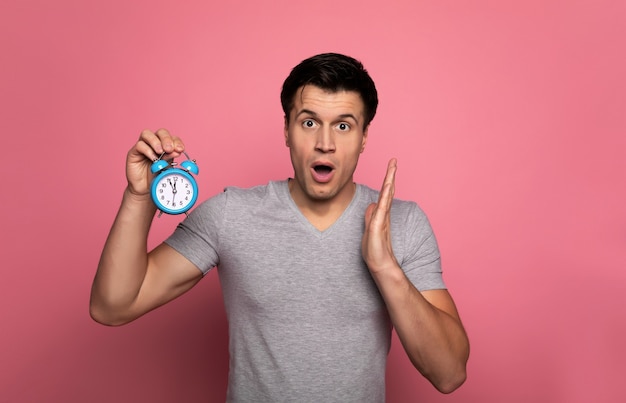 ¡Voy tarde! joven asustado en ropa casual, que sostiene un reloj despertador en su mano derecha y mira a la cámara.