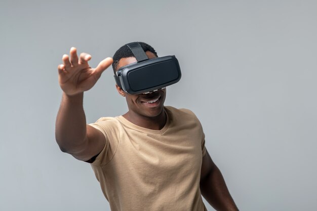 VR-Brille. Lächelnder Afroamerikanermann, der virtuelle Brille im hellen T-Shirt trägt, das seine Hände auf grauem Hintergrund winkt