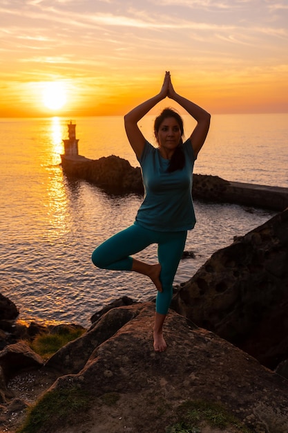 Vrksasana uma mulher fazendo exercícios de meditação e ioga em uma rocha ao pôr do sol ao lado de um farol no mar vida saudável e naturista