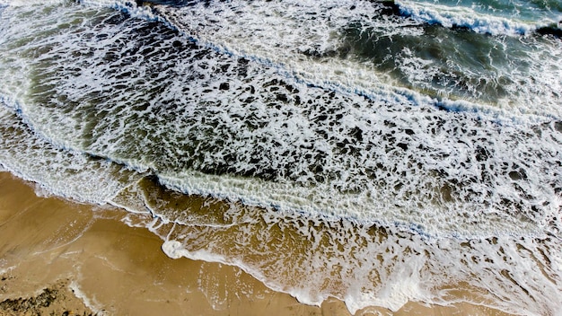 Vuelo aéreo con drones sobre las olas del mar que ruedan hacia la orilla arenosa