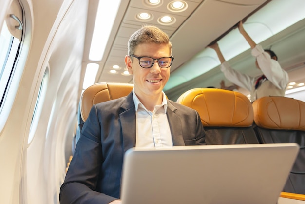 Durante un vuelo, un hombre de negocios con atuendo formal y anteojos mira por la ventana del avión mientras trabaja en una computadora portátil Servicio con acceso a Internet a bordo y un concepto de viaje de negocios