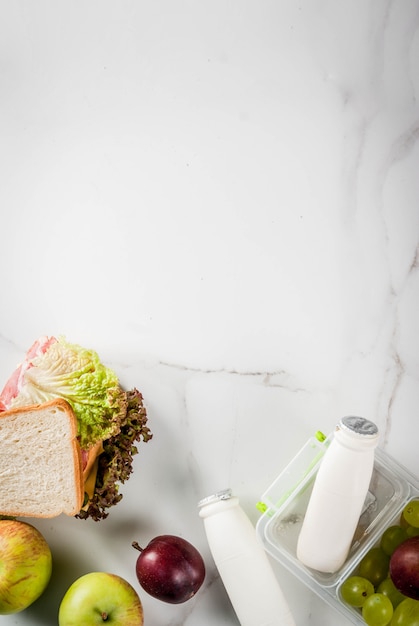 De vuelta a la escuela. Un almuerzo saludable en una caja es fruta fresca, manzanas, ciruelas, uvas, una botella de yogurt y un sándwich con lechuga, tomate, queso y carne. Mesa de mármol blanco. vista superior