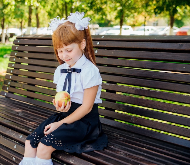 De vuelta a la escuela. Una linda colegiala está sentada en un banco en el patio de la escuela y sostiene una manzana verde en sus manos. Comidas escolares adecuadas para el almuerzo. Una niña va al primer grado.