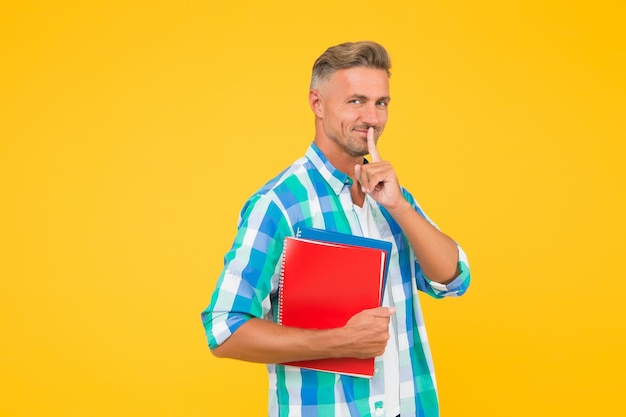 De vuelta a la escuela, el tutor del hombre tiene un libro de ejercicios para la lección. Estudiante universitario con fondo amarillo. Un joven alegre que sostiene un libro y un gesto de silencio sonriente mantiene o revela un copyright secreto