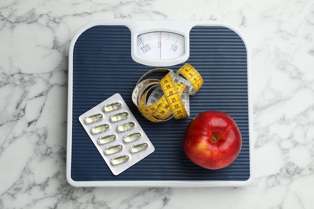 Waage mit Ölkapseln Apfel und Maßband auf weißer Marmortischplatte Ansicht Gewichtsverlust