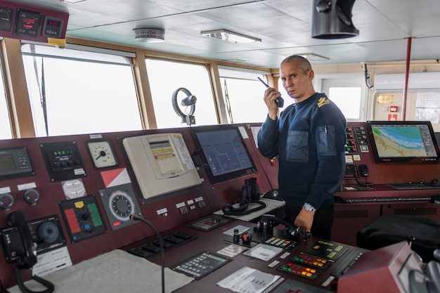 Wachoffizier mit Funk auf der Navigationsbrücke. Kaukasischer Mann im blauen Uniformpullover auf der Brücke eines Frachtschiffs