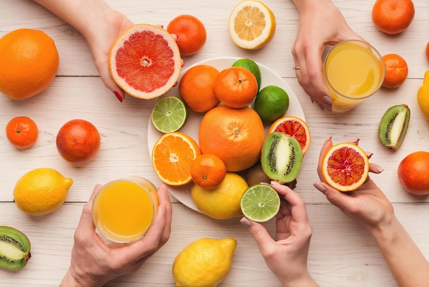 Wählen Sie Ihre Zitrusfrüchte. Viele Hände, die verschiedene Obst- und Saftgläser auf weißem Holzplankenhintergrund halten, Draufsicht