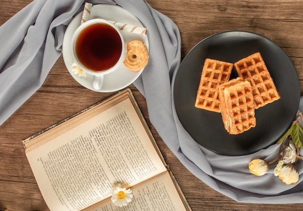 Waffeln auf einer schwarzen Untertasse, eine Tasse Tee und ein Buch. Draufsicht