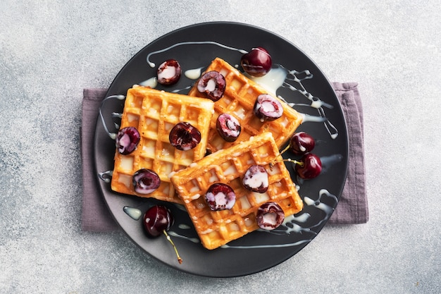 Waffles acanalados belgas con cerezas frescas y crema en un plato. Gofres caseros suaves, postre dulce con cerezas.