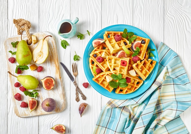 Waffles belgas con bayas frescas, peras, higos en una placa azul sobre una mesa de madera blanca con ingredientes sobre una tabla de cortar, endecha plana, vista horizontal desde arriba