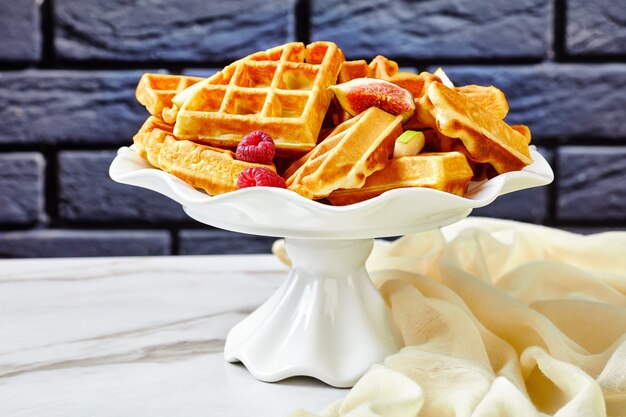 Foto waffles belgas con bayas frescas, peras, higos en un soporte de pastel sobre una mesa de mármol blanco con una pared de ladrillos en la superficie