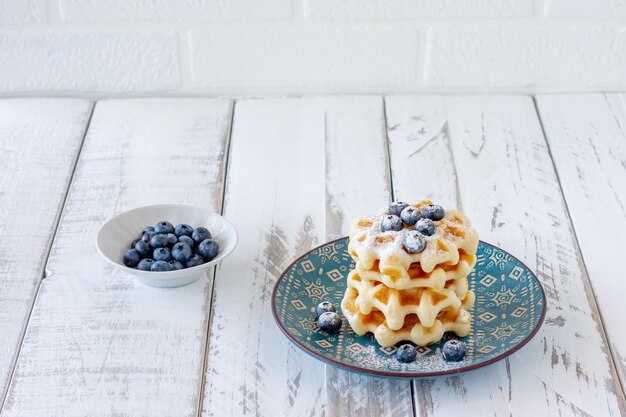 Waffles belgas cuadrados con arándanos y azúcar glas en una placa azul. Sabrosos gofres dulces azucarados.