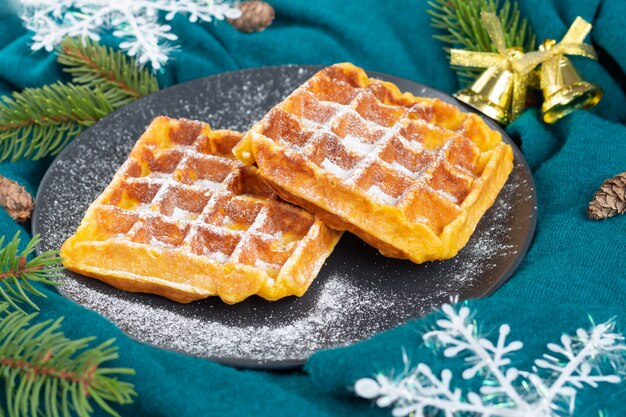 Waffles belgas em um prato preto polvilhado com açúcar de confeiteiro. Clima de Natal e ano novo. Flocos de neve, cones, ramos de pinheiro em tecido turquesa.