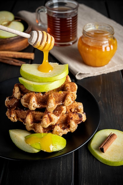 Waffles belgas tradicionais cobertos com fatias de maçã verde e mel descendo do palito e uma xícara de chá. composição do café da manhã na superfície de madeira escura.