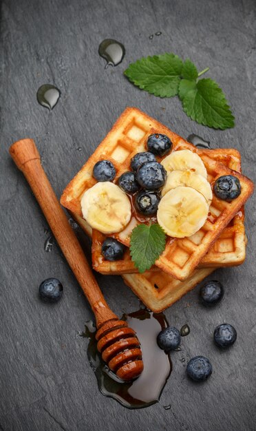 Waffles belgas tradicionales con arándanos, plátano y miel en un plato de pizarra negra, desayuno gourmet,