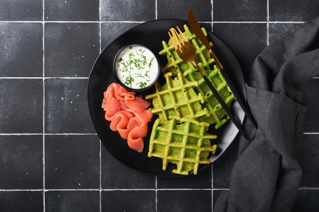 Waffles belgas verdes Espinacas o ajo silvestre o waffles de pesto con salmón rojo y salsa de crema sobre el fondo de la mesa de azulejos negros Delicioso brunch de desayuno en la vista superior de fondo oscuro