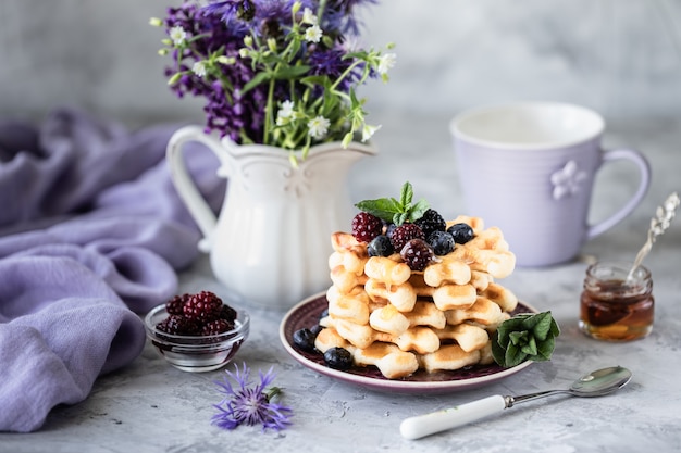 Waffles caseiros com frutas e mel, uma xícara de café na mesa com um buquê de lilases.