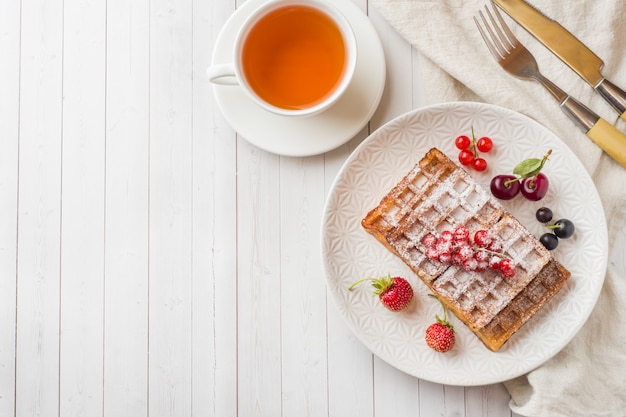 Waffles caseros con bayas de verano en un plato. Una taza de té en una mesa de luz. Enfoque selectivo.