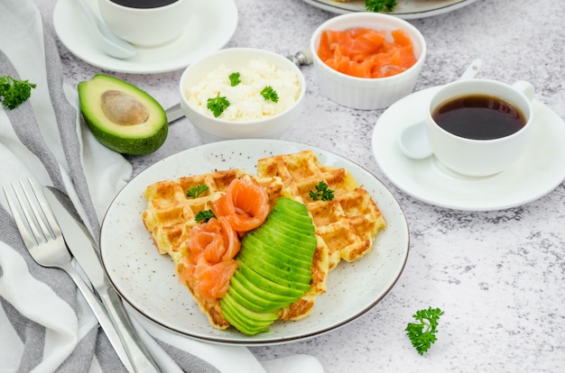 Waffles crocantes de queijo com creme de queijo, salmão defumado e abacate no café da manhã com uma xícara de café sobre um fundo de pedra claro.