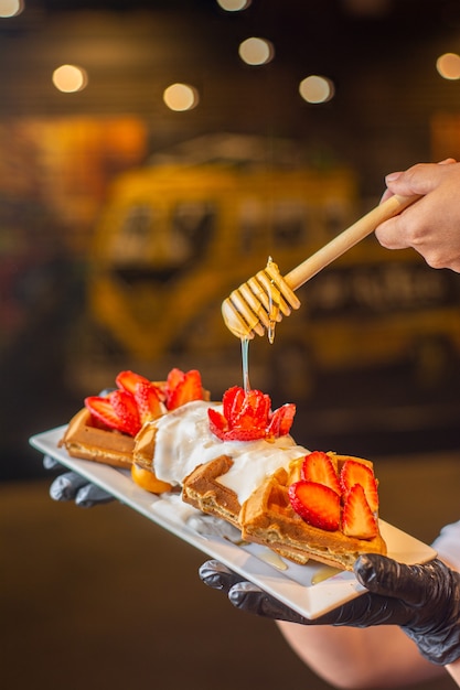 Waffles con fresas y crema batida y miel en la placa blanca en la mano de los camareros