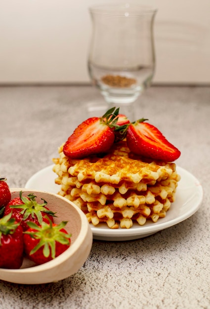 Waffles servidos com morangos em uma tábua branca sobre uma mesa cinza e uma cesta com str maduro