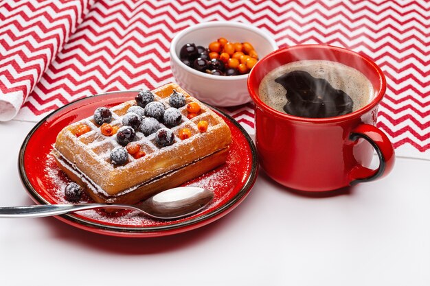 Waffles vienenses polvilhados com pó e decorados com frutas vermelhas. café quente na xícara vermelha.