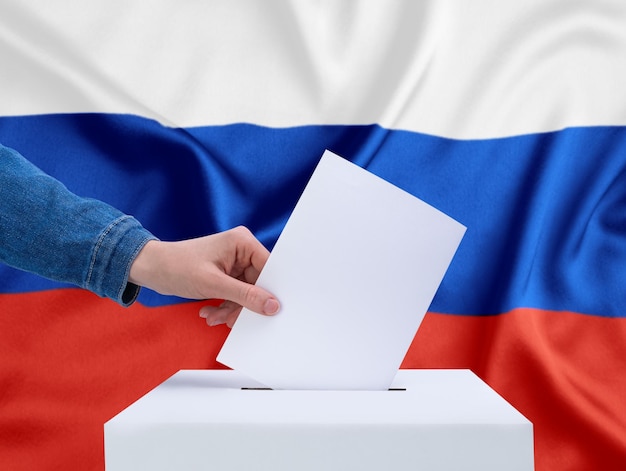 Wahlen in Russland Eine menschliche Hand wirft einen Stimmzettel in die Wahlurne Die russische Flagge im Hintergrund