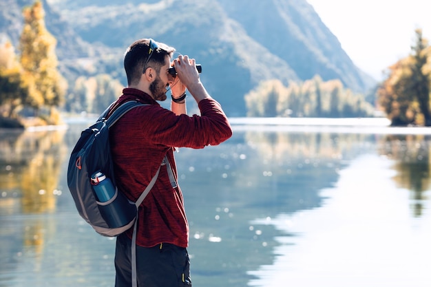 Wanderer junger Mann Reisender mit Rucksack, der die Landschaft mit einem Fernglas im See betrachtet.