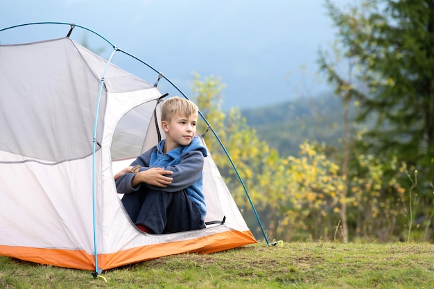 Wanderer-Kind-Junge, der in einem Campingzelt auf dem Bergcampingplatz sitzt und den Blick auf die Sommernatur genießt.