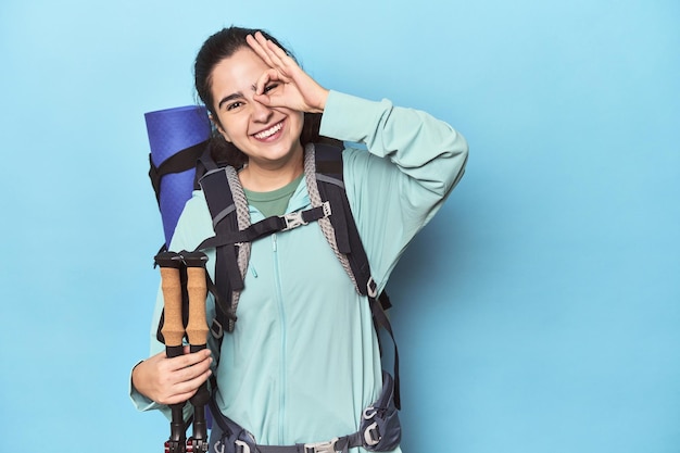 Wanderer mit Rucksack und Stöcken auf blauem Grund, aufgeregt, die Geste „OK“ im Auge behaltend