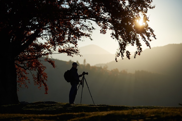 Wanderer touristischer Mann mit Kamera auf grasbewachsenem Tal auf dem Hintergrund der Berglandschaft unter großem Baum.