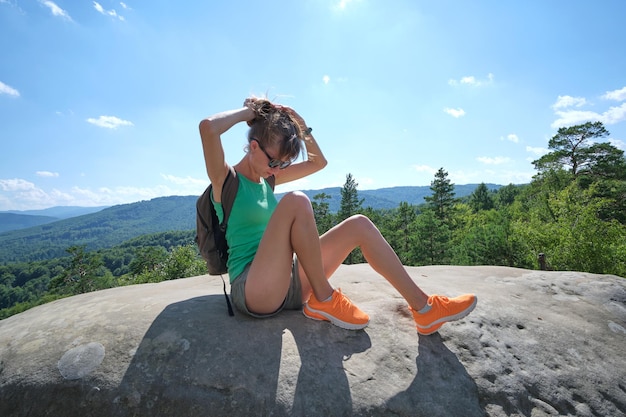 Wanderfrau ruht sich auf einem felsigen Berggipfel aus und genießt die Natur während ihrer Reise auf einem Wildnispfad. Einsame Reisende überquert an heißen Sommertagen eine Route auf einem hohen Hügel. Konzept für einen gesunden Lebensstil