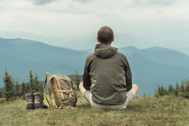 Wandertourist sitzt auf einem Berghügel und blickt auf die Berge mit einem Rucksack und Stiefeln, die neben ihm stehen