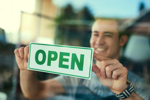 Waren offen Schnappschuss eines jungen Mannes, der das „Geöffnet“-Schild in sein Caféfenster hängt