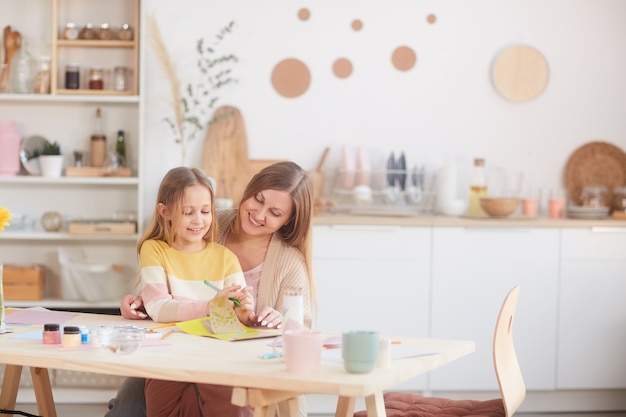 Warm getöntes Porträt der glücklichen Mutter, die kleine Tochter umarmt, während Bilder am hölzernen Küchentisch zeichnen, Raum kopieren