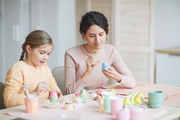 Warm getöntes Porträt der lächelnden jungen Mutter und Tochter, die Ostereier im gemütlichen Kücheninnenraum malen, kopieren Raum